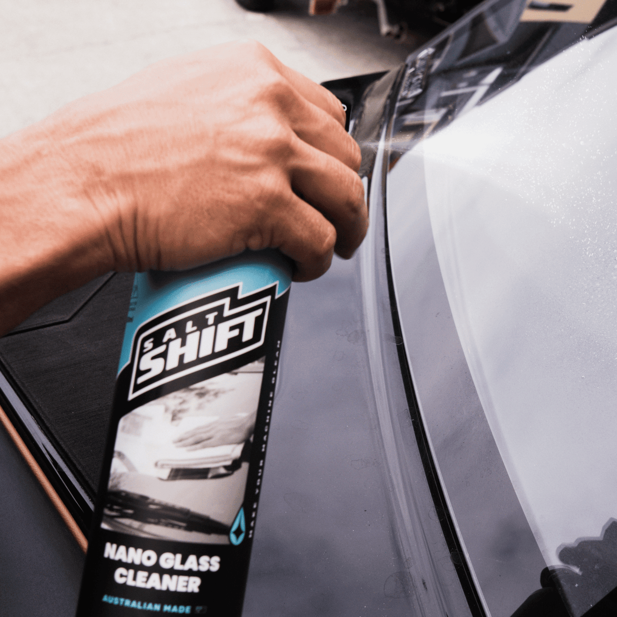 A hand sprays Salt Shift Nano Glass Cleaner on a car windshield for a streak-free shine. Perfect for a crystal-clear view.
