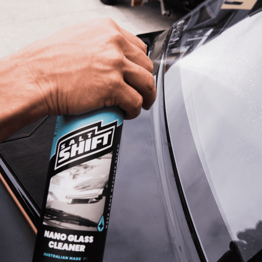 A hand sprays Salt Shift Nano Glass Cleaner on a car windshield for a streak-free shine. Perfect for a crystal-clear view.