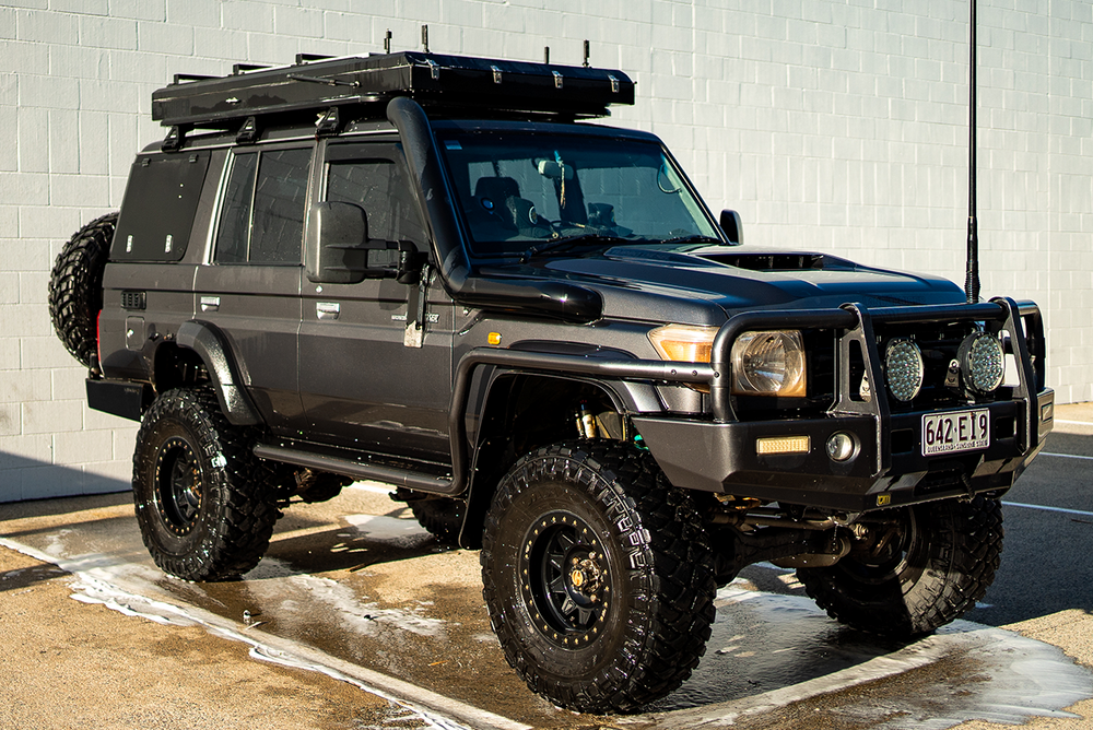 Black off-road vehicle with roof rack parked on a concrete surface against a light gray wall.