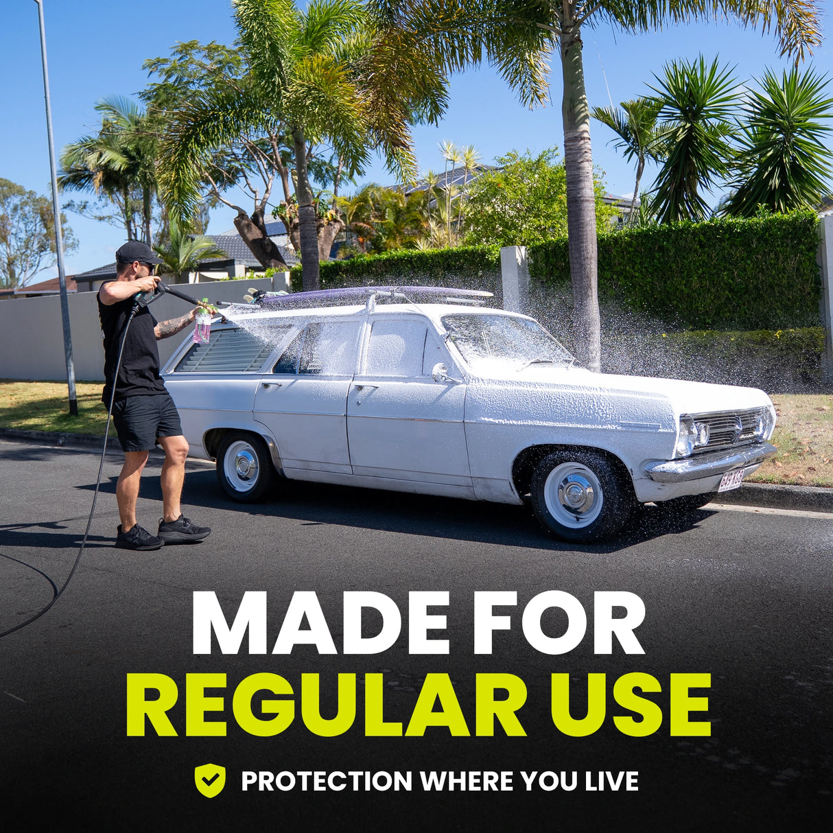 Person washing a white vintage car with palm trees and a clear blue sky in the background.