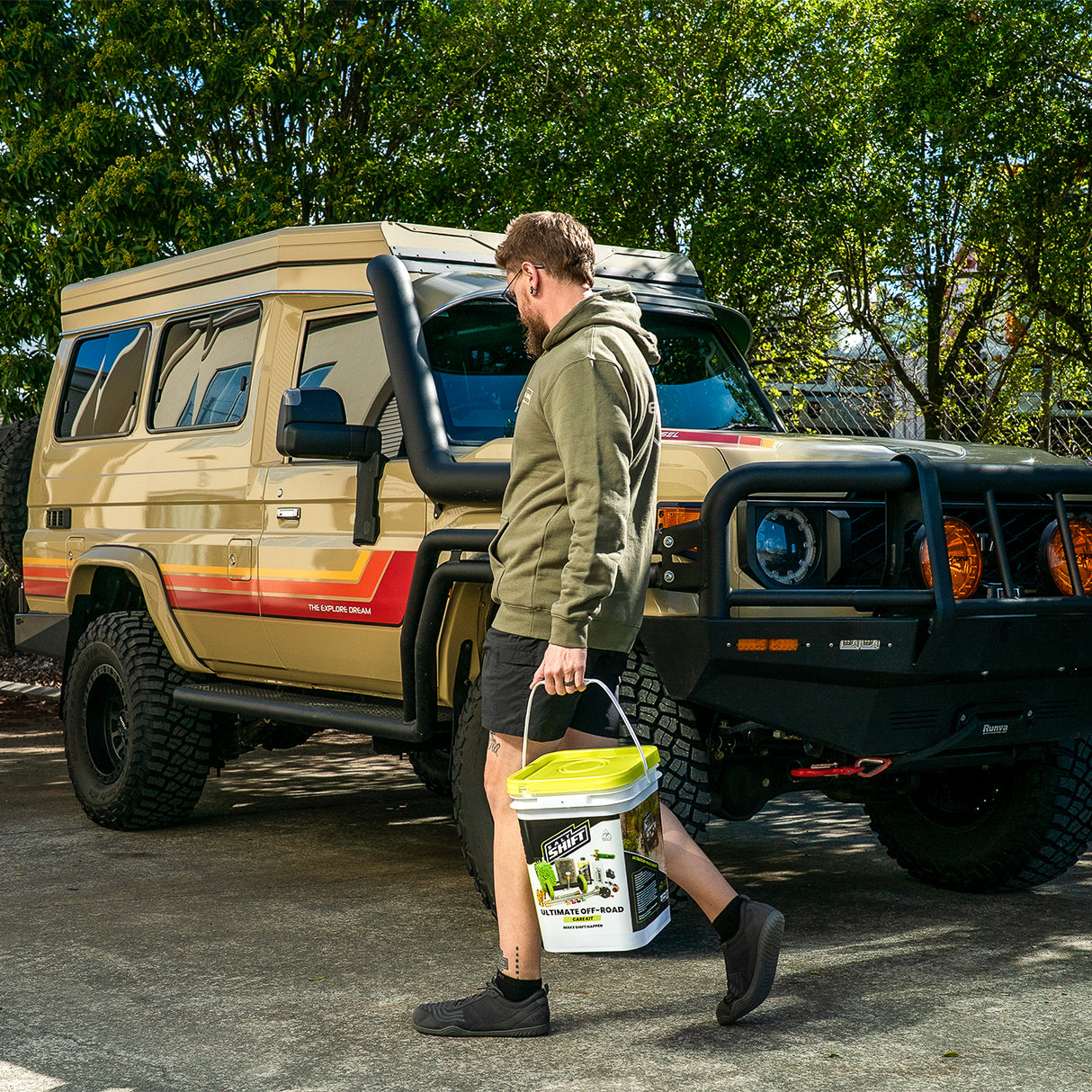 Man standing next to a vintage van holding a bucket, with trees in the background