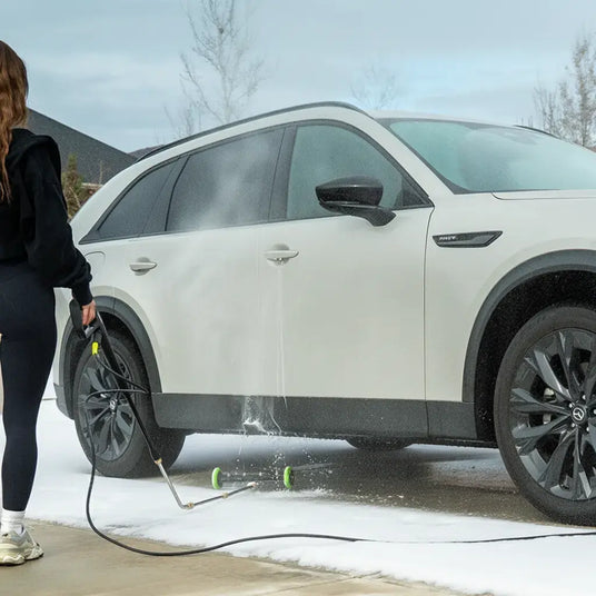 Person using a pressure washer to clean a white SUV in a snowy driveway.