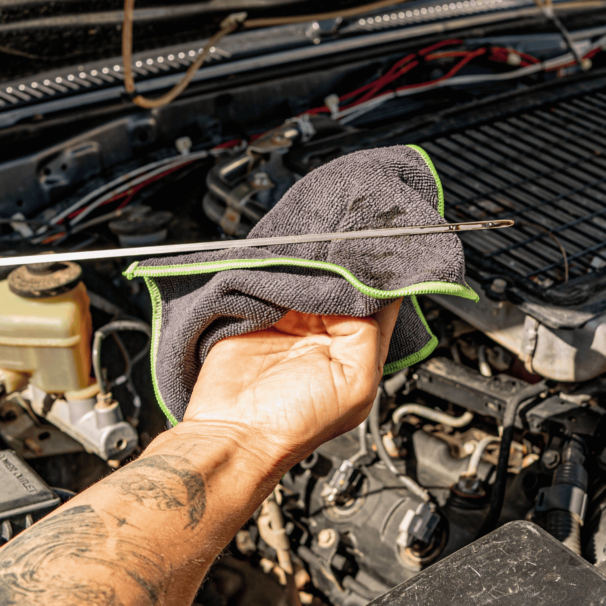 A man holding a black and green microfibre cloth, wiping an engine oil dipstick, with an open engine bay in the background.