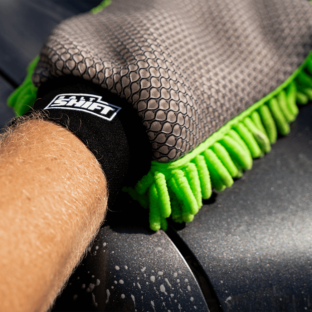 A hand wearing the Salt Shift 2-in-1 Wash Mitt scrubbing a wet car surface. The mitt features a grey hexagonal grip and bright green microfiber strands.