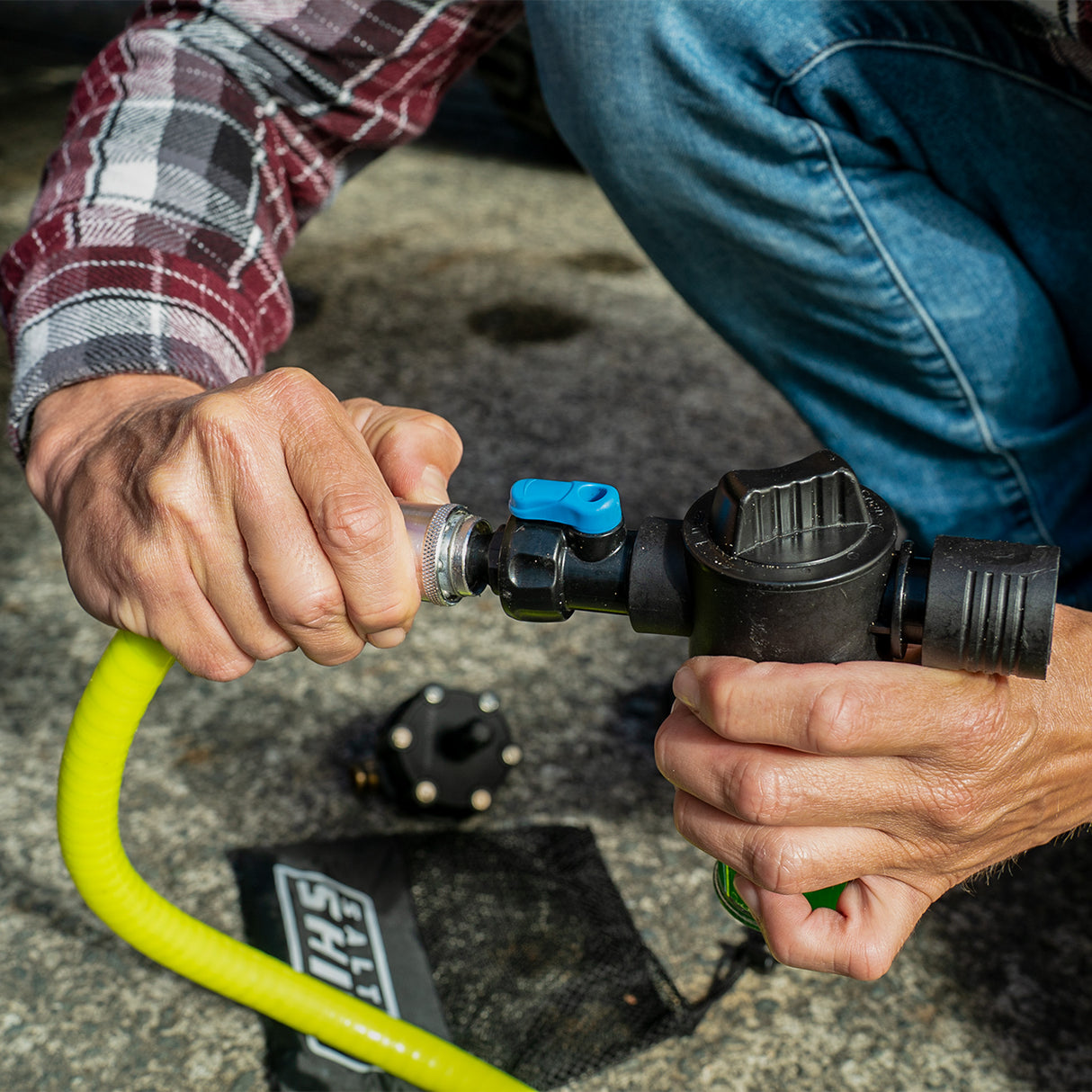 Person connecting a yellow hose to a black water pump with a blue valve on a concrete surface.