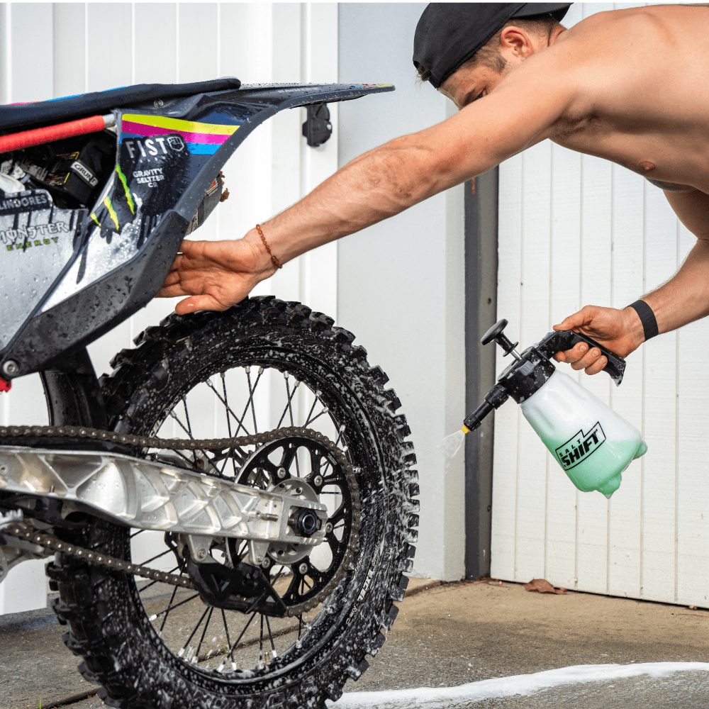 Man using a Salt Shift pump action foamer to clean a dirt bike wheel, removing mud and grime for off-road maintenance.