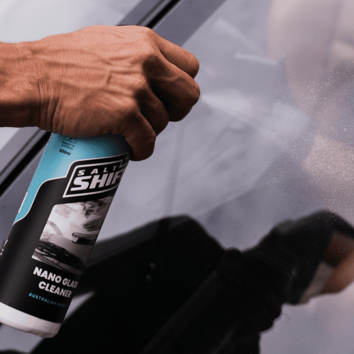 A hand sprays Salt Shift Nano Glass Cleaner on a car windshield for a streak-free shine. Perfect for a crystal-clear view.