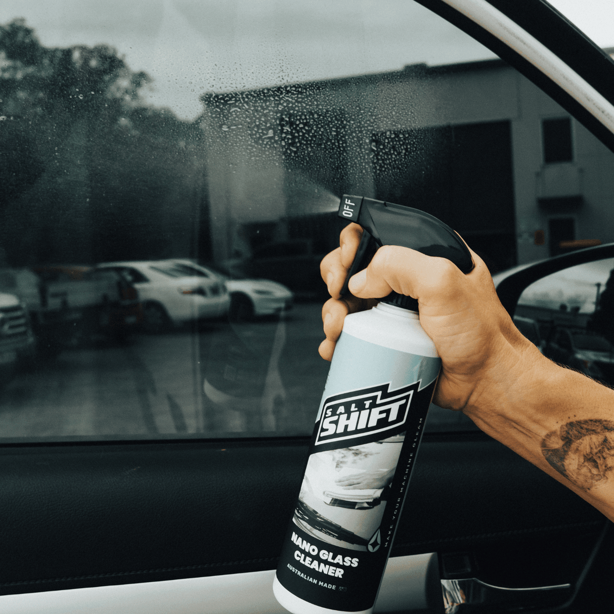Hand spraying Salt Shift Nano Glass Cleaner on a car window to remove dirt and streaks, ensuring a crystal-clear finish. Premium automotive glass cleaning solution.
