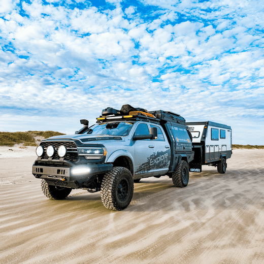 Off-road adventure truck towing a camper trailer on a sandy beach, equipped with gear for exploration and overlanding.