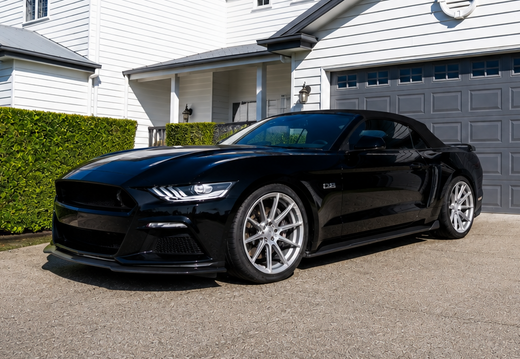 A black Ford Mustang sports car parked on a concrete driveway in front of a modern white house with a gray garage door, green hedges, and a clear blue sky.