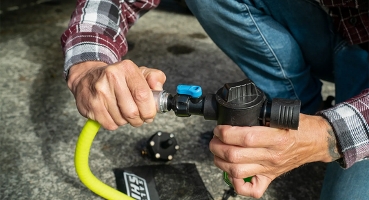 Person connecting a yellow hose to a black water valve on a concrete surface.