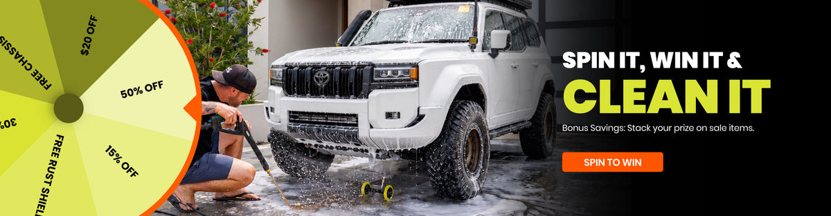 Promotional banner for a car wash service featuring a white SUV being washed, a person kneeling in soapy water, a yellow discount wheel on the left, and text reading 'SPIN IT, WIN IT & CLEAN IT' on a black background.