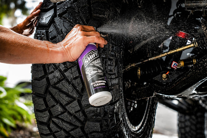 A person sprays tire cleaner from a purple and black can onto a black off-road tire, with the tire and vehicle suspension visible in an outdoor setting.