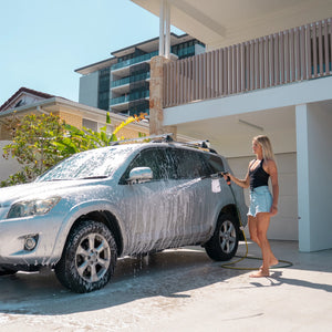 Woman washing a silver SUV with a hose in a driveway.