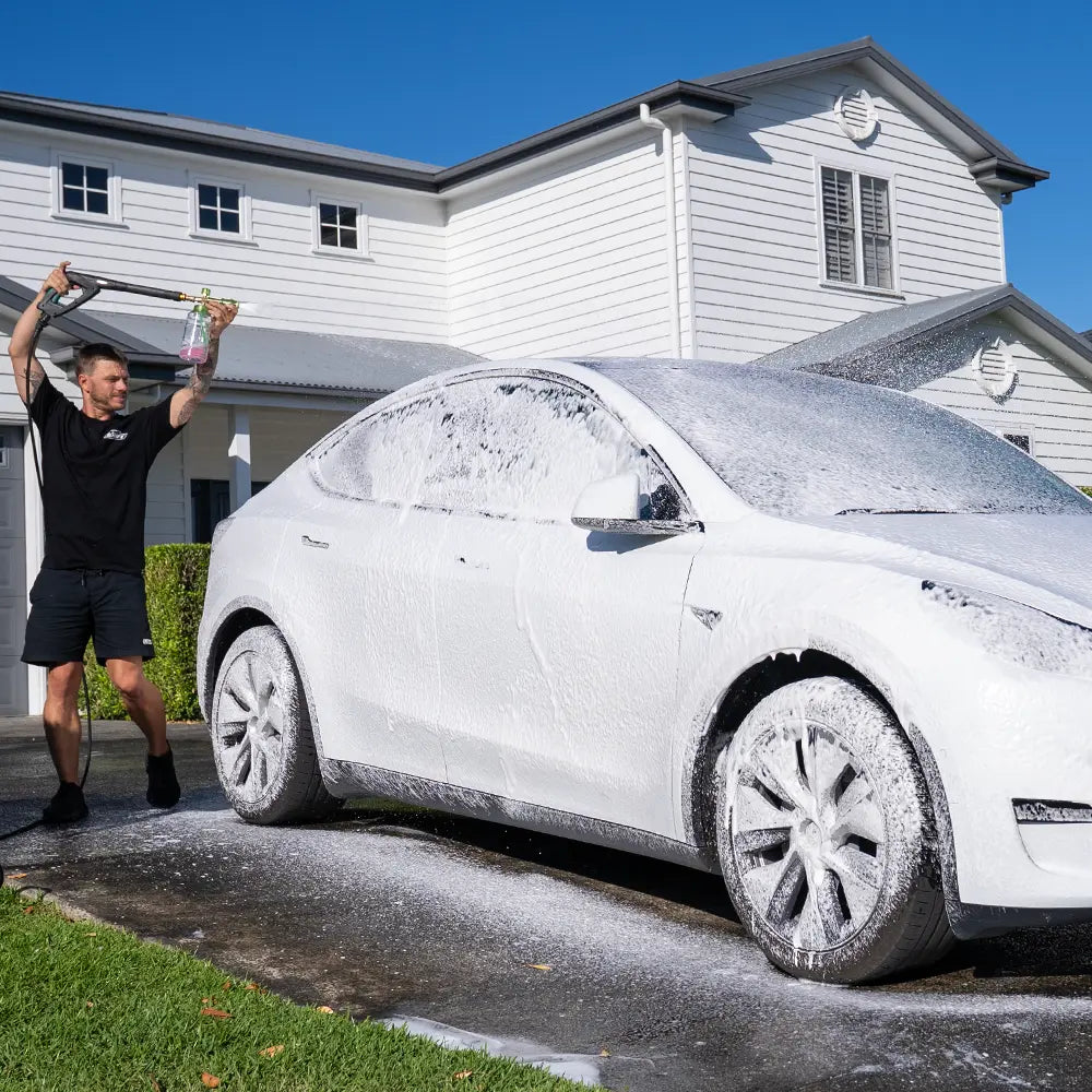 Person washing a white Tesla Model Y with a soapy sponge in front of a house.
