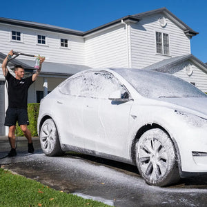 Person washing a white Tesla Model Y with a soapy sponge in front of a house.