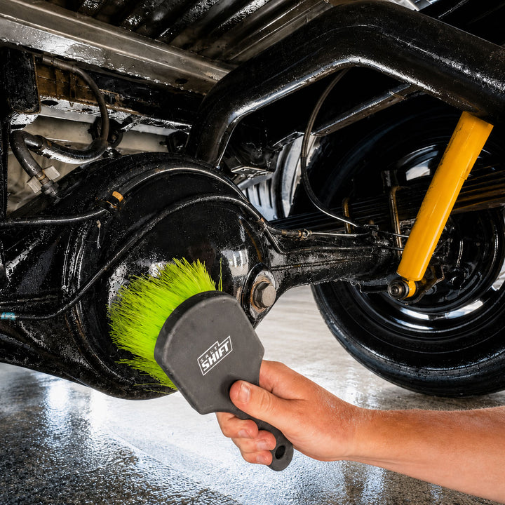 A close-up of a hand using a black and gray brush with bright green bristles to clean the underside of a vehicle near the wheel and suspension components.