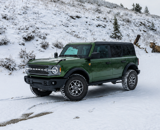 A dark green Ford Bronco SUV is shown driving on a snow-covered mountain road, with snowy hills and evergreen trees in the background under an overcast sky.