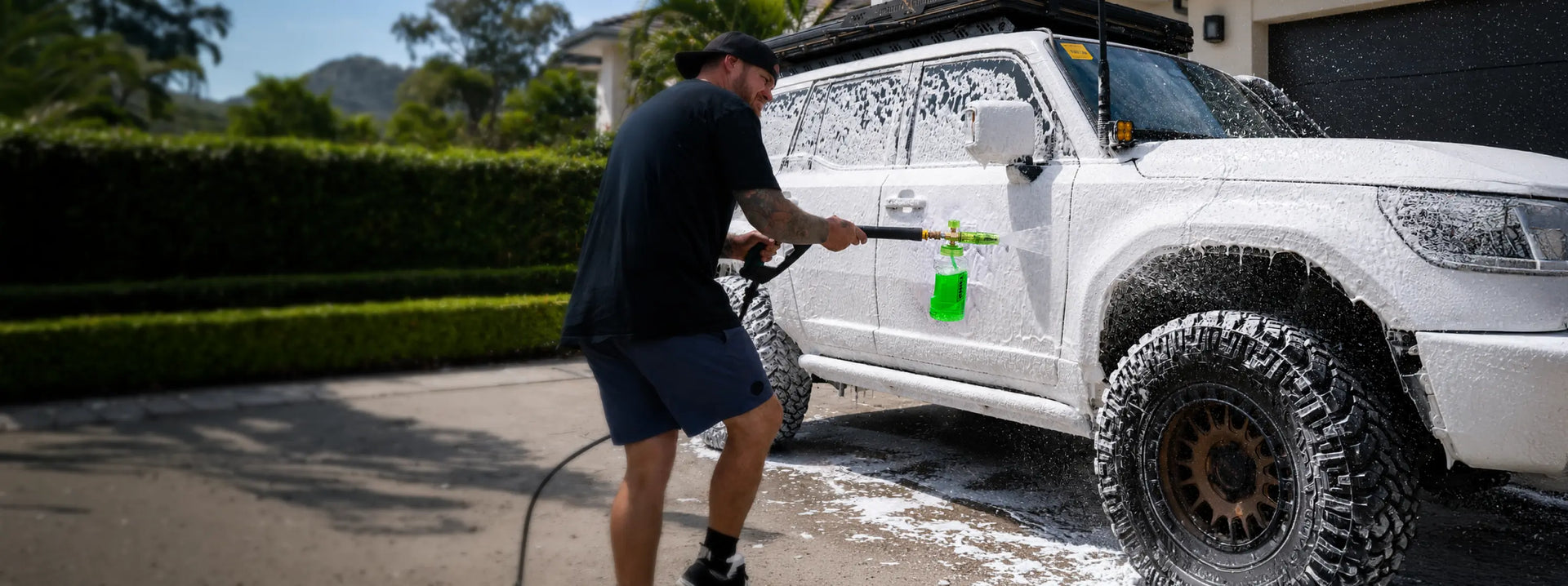 A man in a black t-shirt and shorts washes a white off-road SUV with foam on a driveway in front of a house.