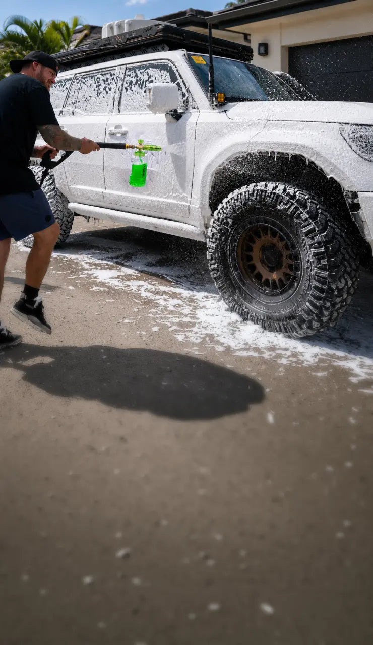 A man washes a white SUV covered in soap foam with a pressure washer in an outdoor driveway setting.