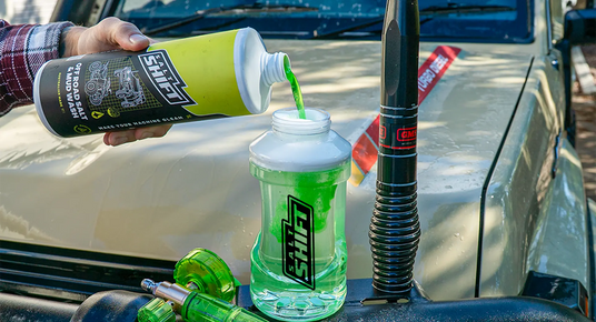 Person pouring a green liquid from a bottle into a clear container with a black logo on a car hood.