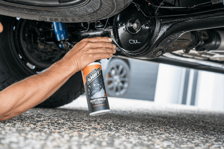 A person's hand holds a can of Salt Shift Rust Shield rust inhibitor spray under a vehicle, with the black metal suspension components and gravel ground visible.