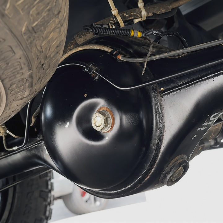 Close-up of a black metal vehicle component, likely a suspension or exhaust part, photographed in a workshop setting with tires and wiring visible.