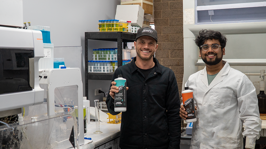 Two individuals in a laboratory setting holding containers, with one wearing a black jacket and the other in a white lab coat.